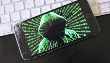 Telefon cu ecran negru, pe cacre e un hacker cu cod verde, pe o tastatură albă, pentru a ilustra virusul periculos care a infectat peste 400 de milioane de telefoane