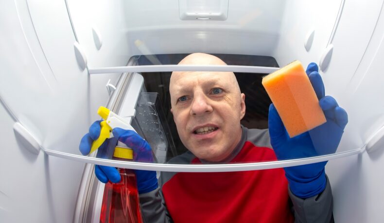 Bărbat chel, îmbrăcat în bluză roșie, cu mănuși albastre, care ține în mâini un burete și o sticlă de curățat, cu capul într-un frigider, pentru a ilustra cum un bărbat a oprit un congelator pentru că îl deranja ce zgomote scotea