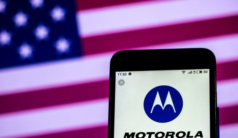 Motorola Solutions, afacerea Motorola care e mai profitabilă, pe ecranul unui telefon, cu steagul american pe fundal