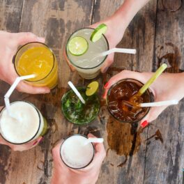 5 persoane care dau noroc cu pahare cu diferite băuturi, fotografiate de sus, cu o masă de lemn sub mâini, pentru a ilustra cel mai bun lichid pentru hidratare