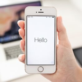 Telefon iPhne alb ținut în mână de utilizator, care are hello pe ecran, cu un laptop pe fundal, pentru a ilustra funcția apple care îți poate imita vocea