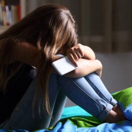 Adolescentă care stă pe pat, cu capul între genunchi, cu telefonul în mână, pentru a ilustra cum o adolescentă a cheltuit toate economiile familiei pe jocuri video