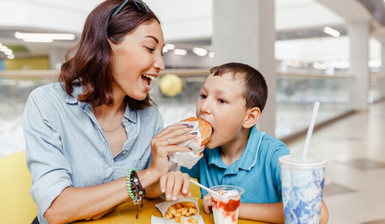 Mamă care își hrănește băiatul cu fast food, un buger. Stau așezaț la masă, cu fast food în fața lor, pentru a ilustra cum a fost descoperită o mutație genetică nemaivăzută