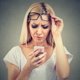 Femeie blondă care își ridică ochelarii pentru a se uita în telefon pentru a ilustra soluția apple pentru cum ne afectează smartphone-urile ochii