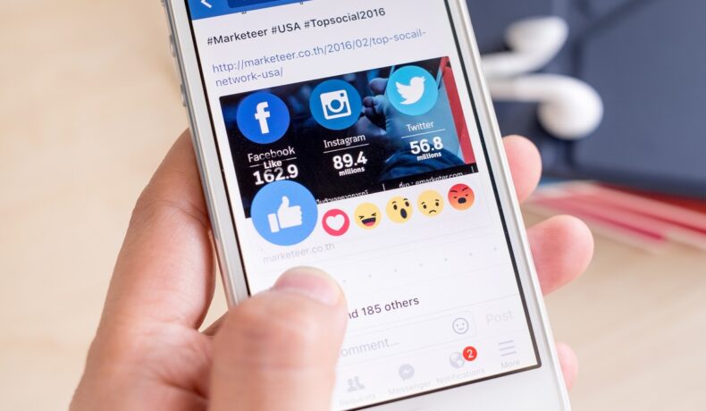 Utilizator care ține un telefon în mână și reacționează la o postare pe facebook cu ajutorul emoji-ului cu degetul mare ridicat