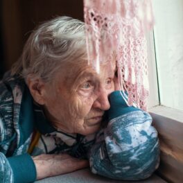 Femeie bătrână, îmbrăcată într-un halat albastru, care stă tristă la o masă și se uită pe fereastră, pentru a ilustra ce efect poate avea singurătatea asupra diabeticilor