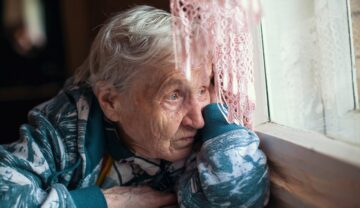 Femeie bătrână, îmbrăcată într-un halat albastru, care stă tristă la o masă și se uită pe fereastră, pentru a ilustra ce efect poate avea singurătatea asupra diabeticilor