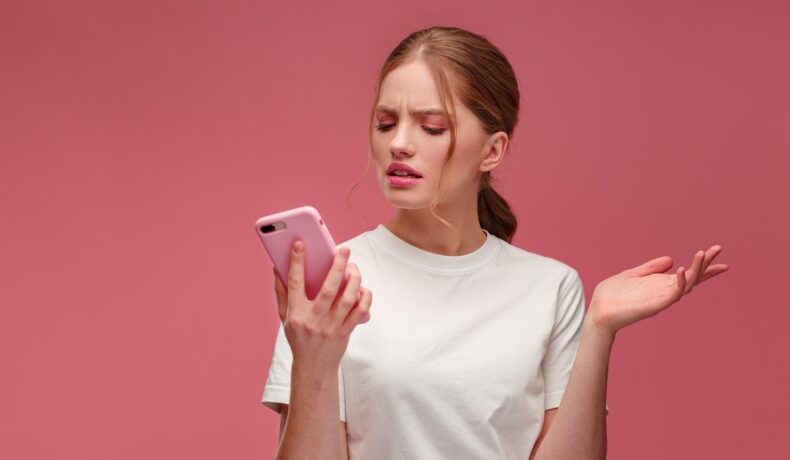 Femeie roșcată care are un telefon roz în mână, pe fundal roz, îmbrăcată în bluză albă, pentru a ilustra de ce merg mai greu telefoanele vara
