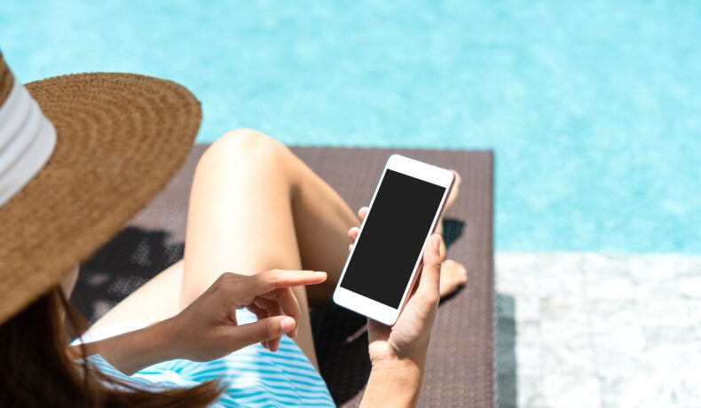 Femeie îmbrăcată în rochie albastră cu pălărie de soare, lângă piscină, care folosește un telefon mobill alb, pentru a ilustra cum telefonul tău poate fi în pericol vara