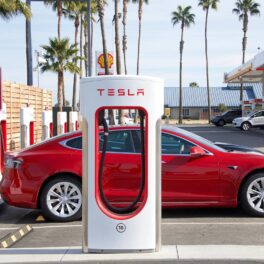 Mașină tesla roșie lângă un încărcător supercharger alb Tesla, pentru a ilustra cum Tesla e acuzată că ar fi indus în eroare consumatorii