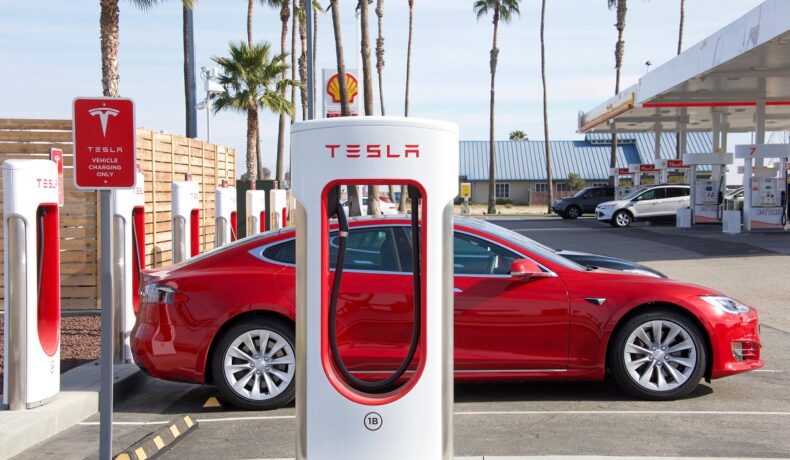 Mașină tesla roșie lângă un încărcător supercharger alb Tesla, pentru a ilustra cum Tesla e acuzată că ar fi indus în eroare consumatorii