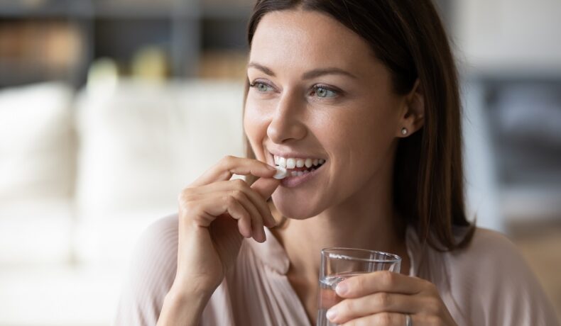 Femeie brunetă care zâmbește și înghite o pastilă, similară cu un medicament comun care tratează hipertensiunea