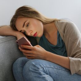 Femeie blondă care stă spprijinită de o canapea gri, cu un telefon în mână, îmbrăcată în bej și blugi, pentru a ilustra ce se întâmplă cu ochii tăi când te uiți la ecranul telefonului