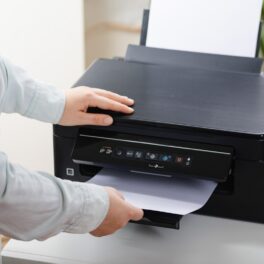 Persoană care folosește o imprimantă neagră și scoate o hârtie din ea, pentru a ilustra ce trebuie să faci cu imprimanta înainte să o arunci