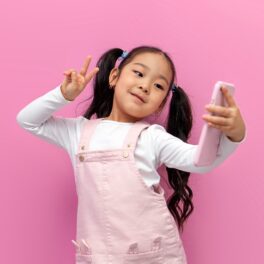 Fetiță asiatică, cu codițe și părul brunet, îmbrăcată în alb și roz, pe fundal roz, cu un telefon roz în mână, pentru a ilustra cum China recomandă limitarea timpului petrecut pe smartphone