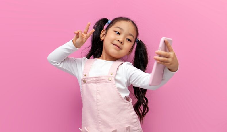 Fetiță asiatică, cu codițe și părul brunet, îmbrăcată în alb și roz, pe fundal roz, cu un telefon roz în mână, pentru a ilustra cum China recomandă limitarea timpului petrecut pe smartphone