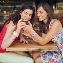 2 femei care stau la masă și se uită la un telefon, una dintre ele e îmbrăcată în rochie, cealaltă poartă antaloni și bluză roșie, pentru a ilustra singurul cuvânt care se pronunță la fel în toate limbile