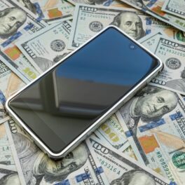 Telefon cu ecran negru și carcasă albă pe un maldăr de dolari, pentru a ilustra cele mai vândute telefoane din 2023