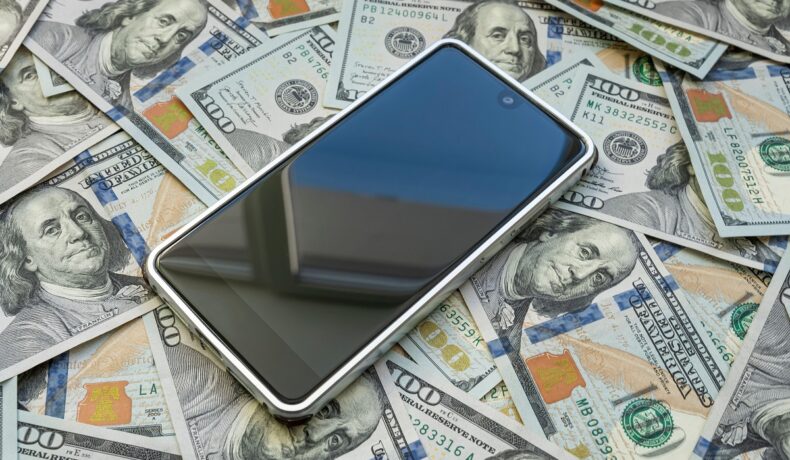 Telefon cu ecran negru și carcasă albă pe un maldăr de dolari, pentru a ilustra cele mai vândute telefoane din 2023