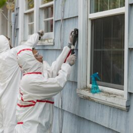 2 meșteri îmbrăcați în costume de rotecție albe care analizează o casă albastră pentru plumb, metalul care e mai periculos decât fumatul