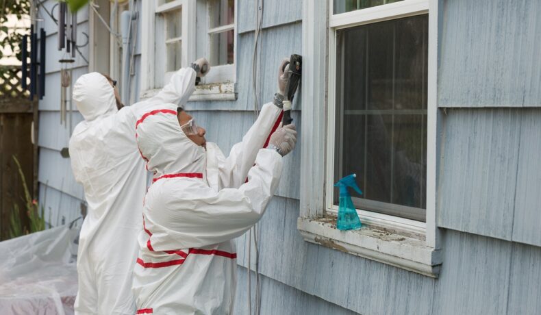 2 meșteri îmbrăcați în costume de rotecție albe care analizează o casă albastră pentru plumb, metalul care e mai periculos decât fumatul