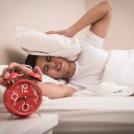 Persoană care stă în pat, cu o pernă pe cap, care închide alarma de pe un ceas roșu, pentru a ilustra de ce e bine să apeși pe snooze