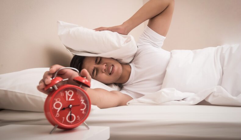 Persoană care stă în pat, cu o pernă pe cap, care închide alarma de pe un ceas roșu, pentru a ilustra de ce e bine să apeși pe snooze