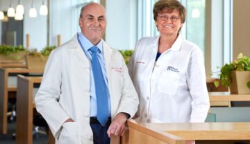 Katalin Kariko şi Drew Weissman, îmbrăcați în halate albe de cercetători, lângă un birou din lemn, ce au câștigat Premiul Nobel pentru medicină în 2023