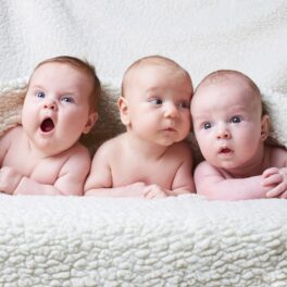 3 bebeluși care stau pe un pat, cu o pătură albă pufoasă, pentru a ilustra luna în care se nasc cei mai mulți copii