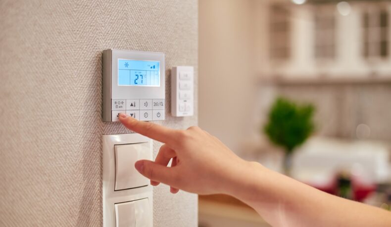 Mână de femeie care ajustează un termostat alb, pe perete, cu temperatura optimă pentru termostat