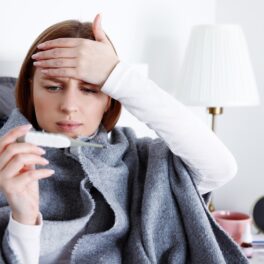Femeie bolnavă, care s eține de frunte și se uită la un termometru, pe fundal alb, pentru a ilustra ce se întâmplă când ai febră