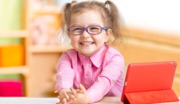 Fetiță mică și blondă, similară cu cea mai tânără membră Mensa, cu o cămașă roz, la un birou alb, cu ochelari