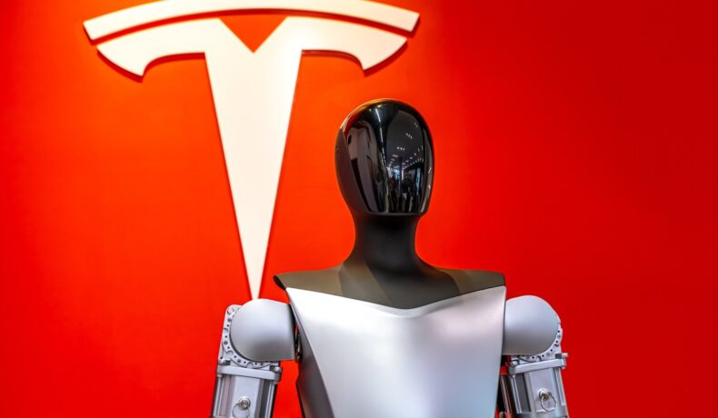 Robot Optimus, care stă în fața unui logo Tesla roșu, pentru a ilustra cum Tesla a dezvăluit Optimus Gen 2