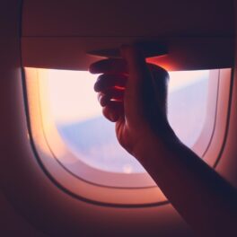 Mână care trage oblonul unui geam de avion, pentru a ilustra de ce trebuie să ridici obloanele geamurilor avionului la decolare și aterizare