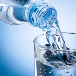 Sticlă de apă din plastic, turnată într-un pahar, pe fundal albastru, pentru a ilustra pericolul ascuns din apa îmbuteliată