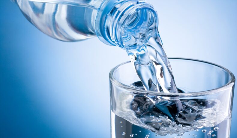 Sticlă de apă din plastic, turnată într-un pahar, pe fundal albastru, pentru a ilustra pericolul ascuns din apa îmbuteliată