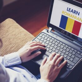 Persoană care ține un laptop pe genunchi, cu o imagine learn romanian pe ecran și steagul României, pentru a ilustra cât de grea e limba română